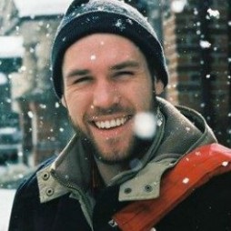 Portrait of smiling man with light skin and brown beard and mustache, wearing a beanie, undercoat and outer coat. Snow falls down around him.