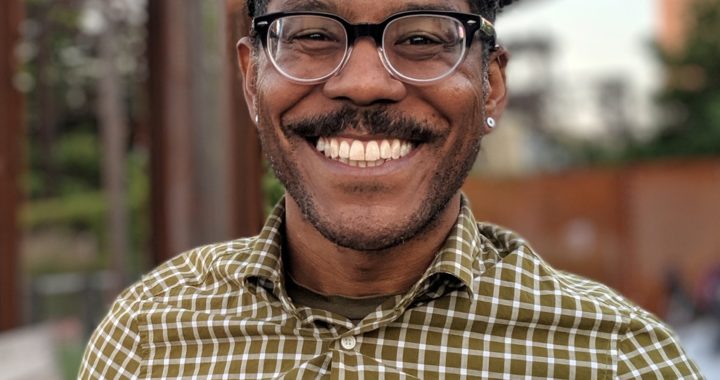 Portrait of a Black man with glasses, mustache and beaming smile wearing a plaid shirt.