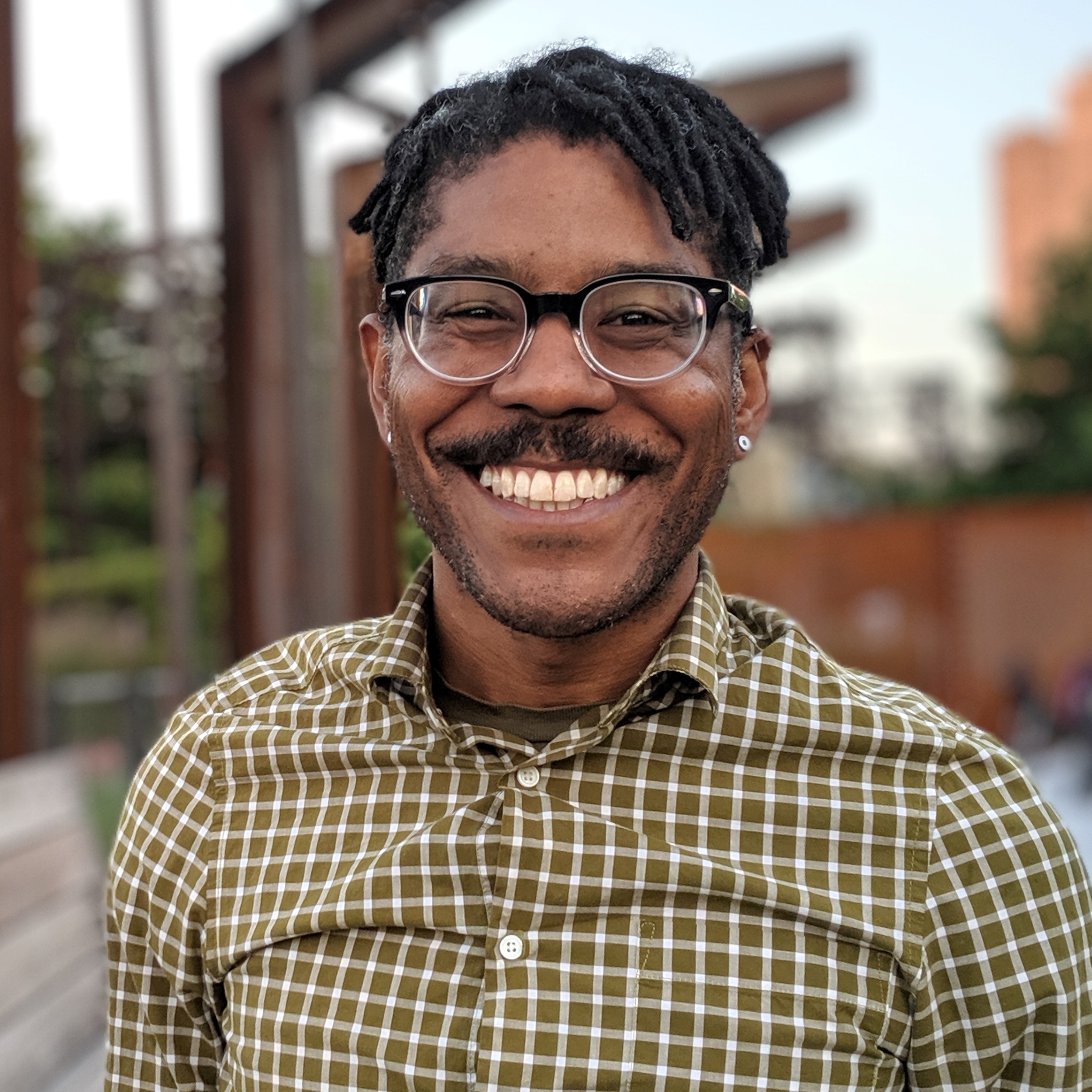 Portrait of a Black man with glasses, mustache and beaming smile wearing a plaid shirt.