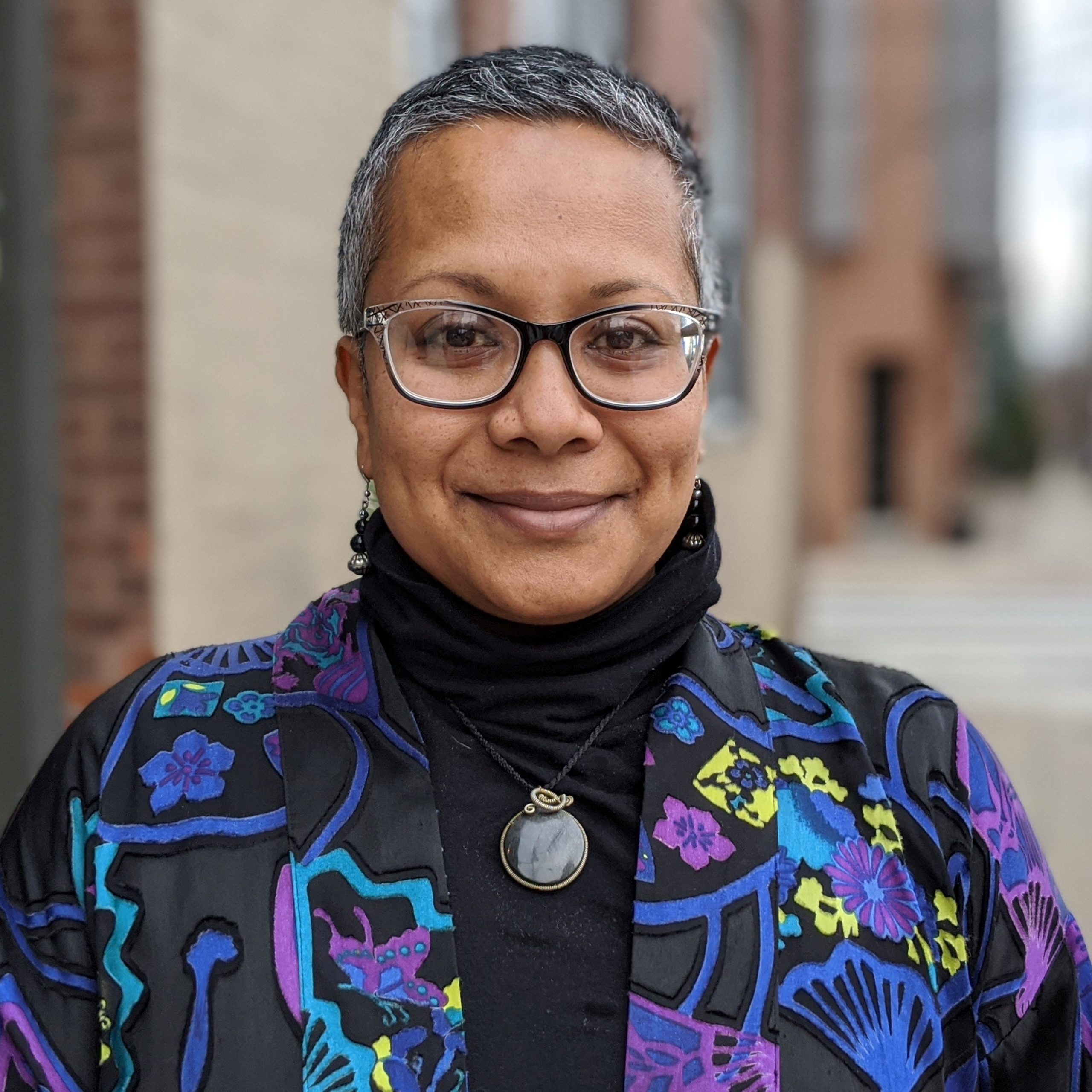 Portrait of person with dark skin, salt-and-pepper hair cropped short, wearing glasses, a blue, purple and black patterned shirt over a black turtleneck with a medallion necklace.