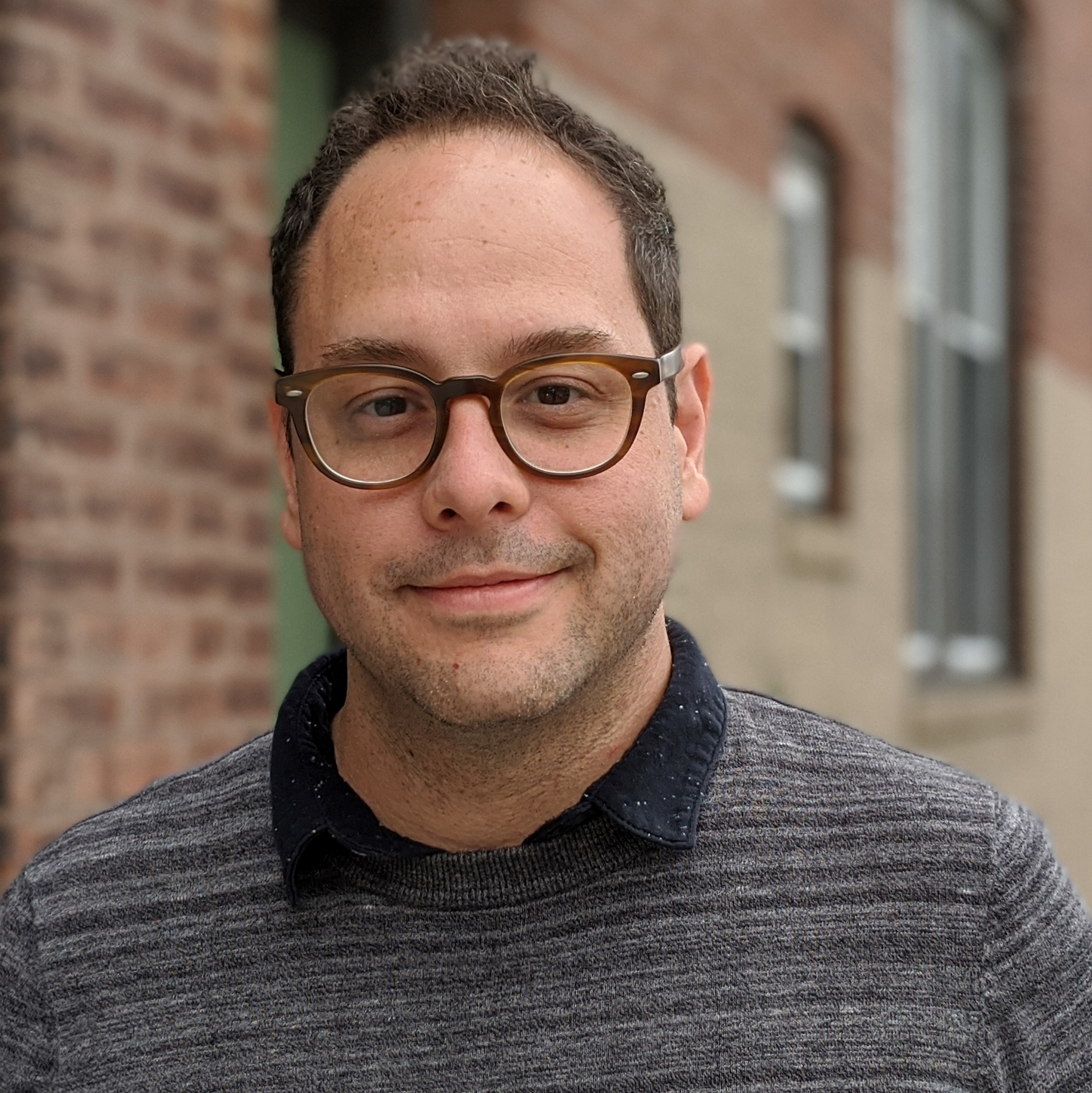 Portrait of man with light skin, short hair and glasses wearing a polo shirt and a slight grin.