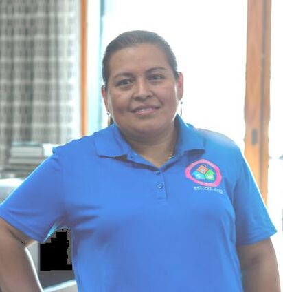 Portrait of smiling person with dark brown skin and brown hair and dangling earrings standing with one hand on hip. The person is wearing a blue short-sleeved polo shirt with a red, blue and green logo on it.