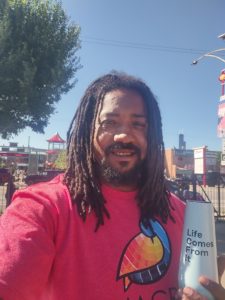 Person with dark brown skin, beard, mustache and long locs smiles while holding up a water bottle that says "Life comes from it". The person wears a light red shirt with a colorful bird decal.