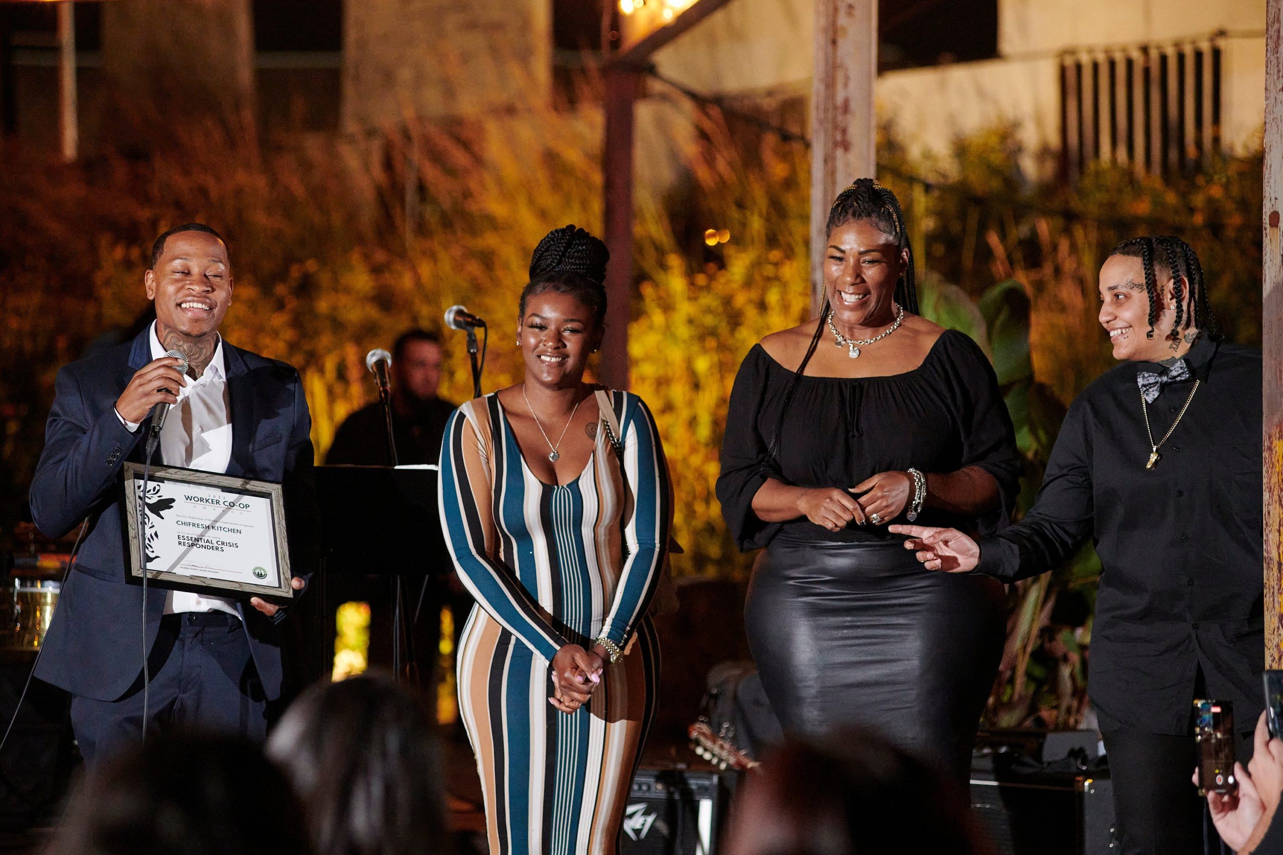 A photo of four worker-owners of ChiFresh Kitchen standing on a stage with microphones in the background. The person on the left wears a white collared shirt and a navy suit, holds a certificate and a microphone and smiles into it. The second coworker on the left wears a long sleeve blue striped dress and their hair in a bun of braids and crosses their arms and smiles. The third coworker on the right wears a black blouse and leather skirt and the coworker on the right wears short black braids, a black collared shirt and a bowtie and a gold chain and points and smiles to their coworker with the microphone.