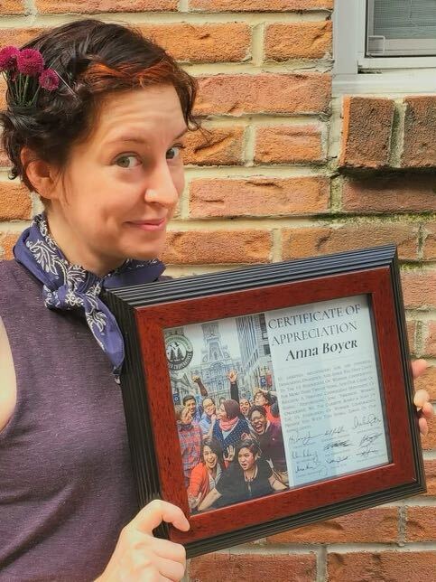 A person with white skin and short brown hair with orange highlights smiles and poses playfully while holding up a certificate of appreciation.