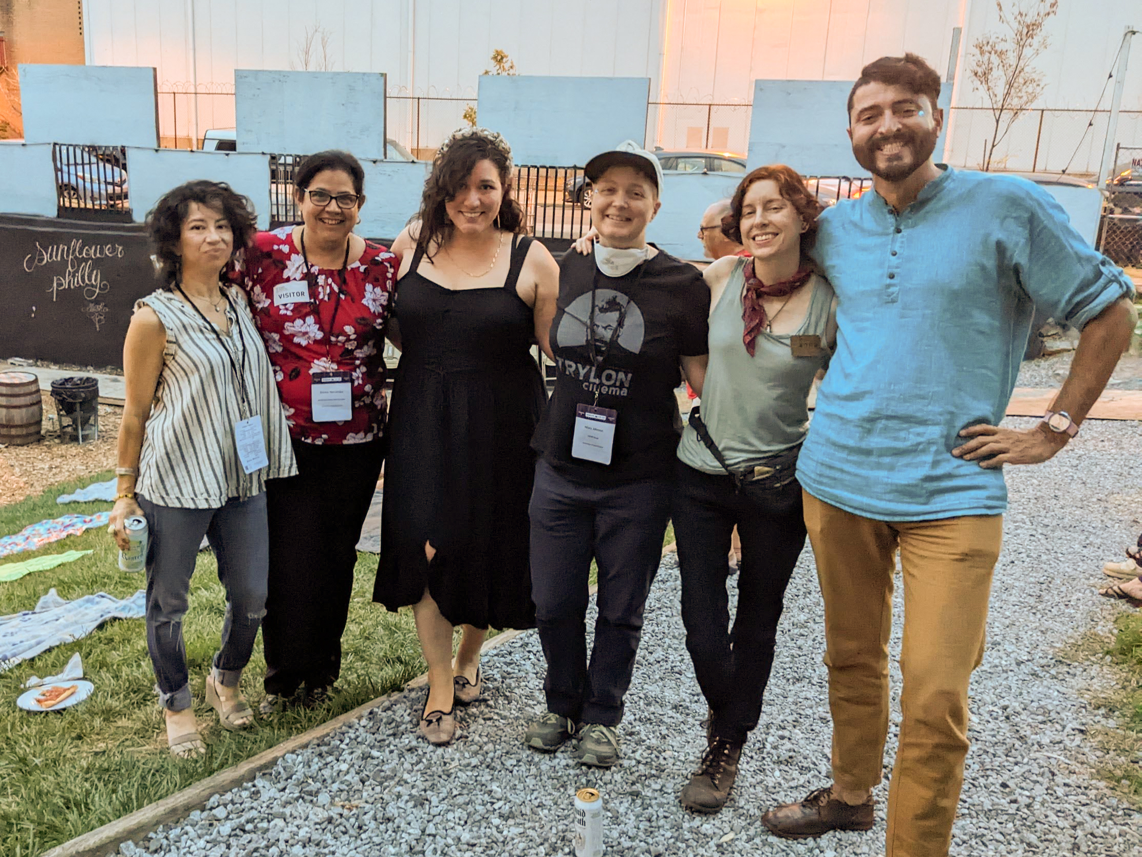 A group of six people with skin tones ranging from pale white to brown pose arm-in-arm while smiling joyfully at an outside social event.