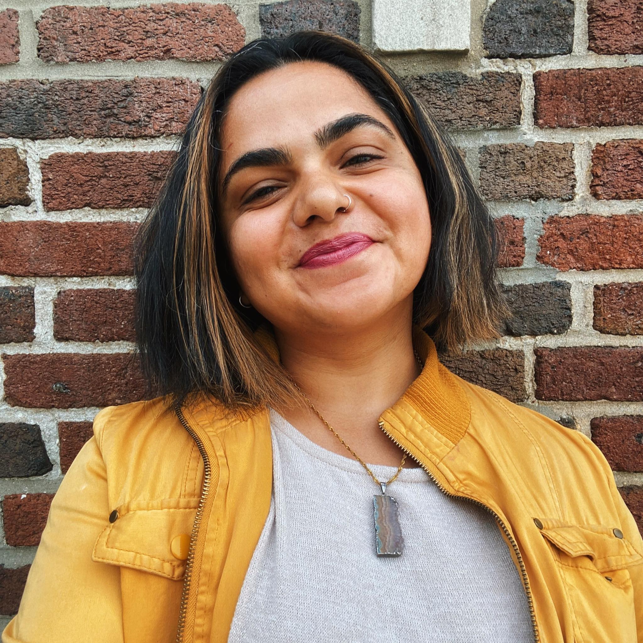 Tehmina Portrait of a person with a warm smile, brown skin, shoulder length brown hair parted down the middle wearing a light grey tee shirt with a glossy yellow leather fabric blend jacket standing in front of a brick wall background.