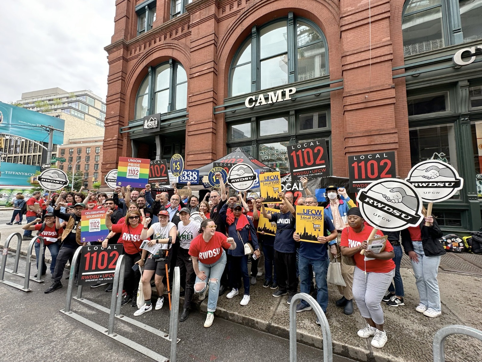Una foto grupal de mas de 20 personas trabajadoras con carteles manifestando frente un edificio de ladrillos en nueva york.