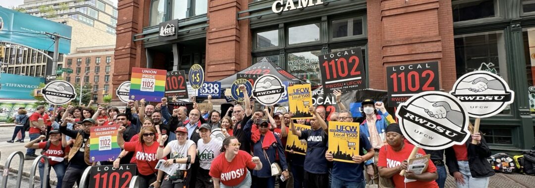 A group of more than 20 people with signs and posters pose in front of a brick building at a worker rally.