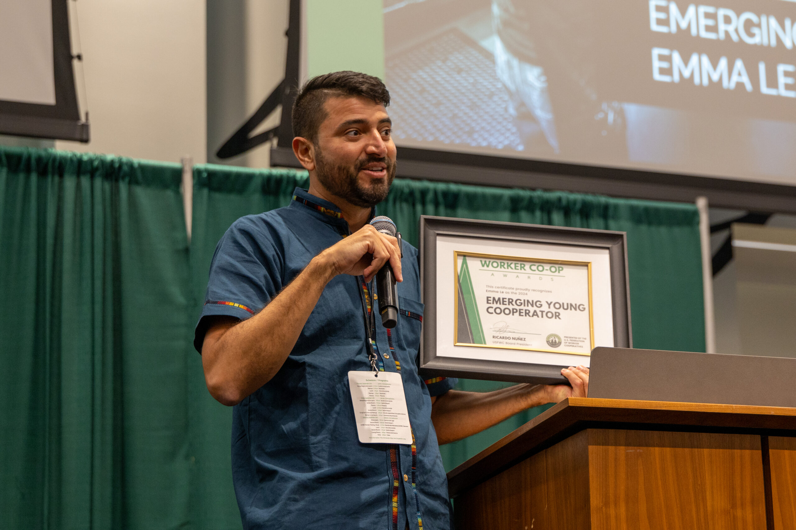 A photo of a tall person with short brown hair and a short beard wearing a blue shirt holding a microphone and a framed award certificate with text that reads worker co-op awards emerging young cooperator