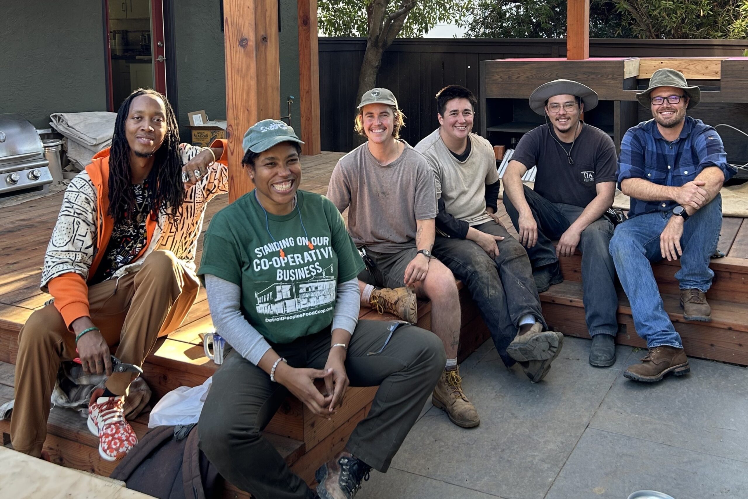 Photo of worker members of Root Volume and Repaired Nations sit on a wooden deck and under a wooden pergola smiling at the camera. In the front is Sabine Dabady, who has brown skin and short hair and wears a long sleeve shirt that reads "standing on our cooperative business" and wears a baseball cap.