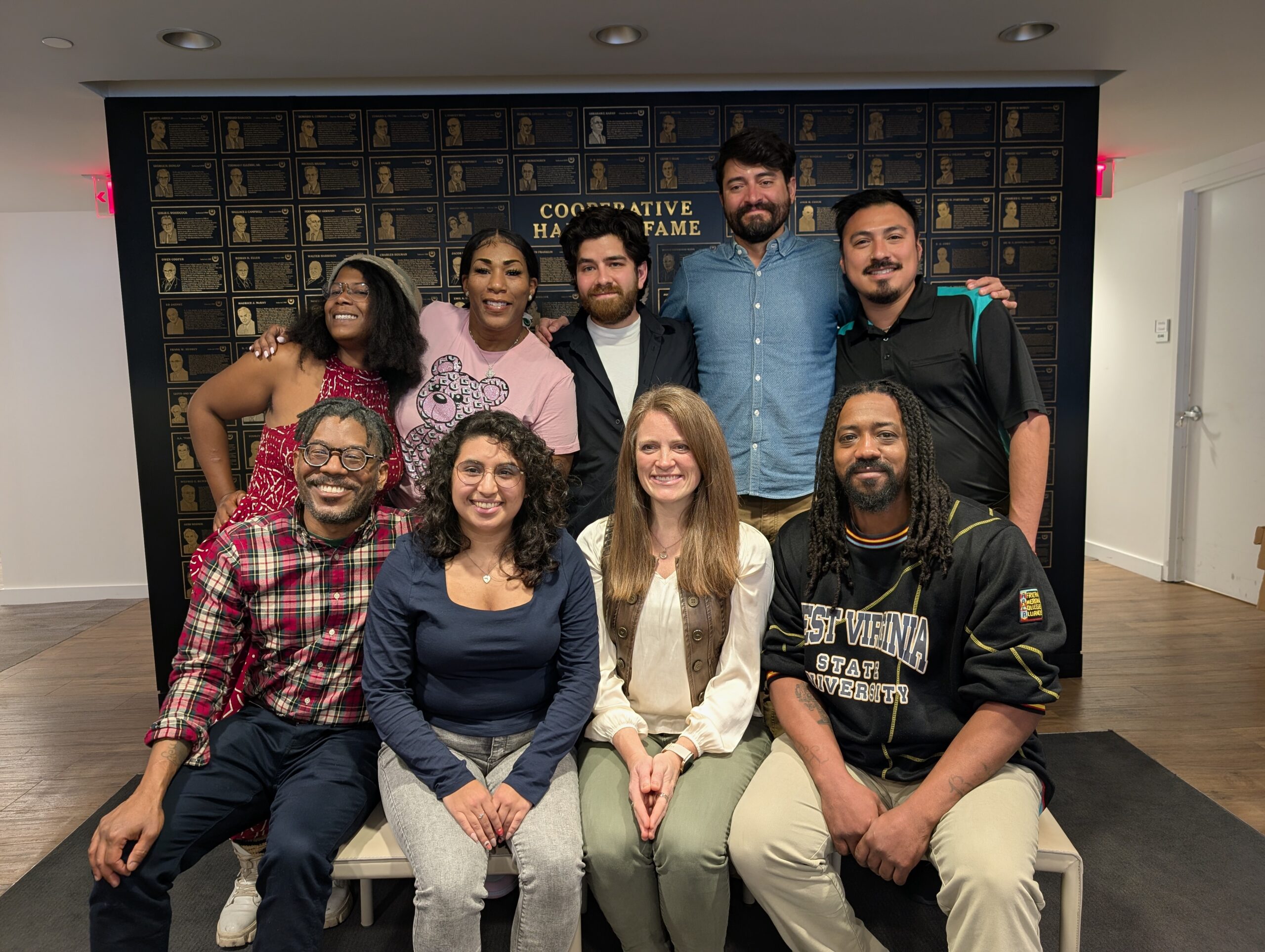 Group of nine people of different races and genders posing together and smiling joyfully in a warmly lit room with bronze wall background