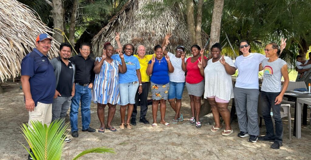 A group of over ten people stand together and smile on the beach with palm tree sin the background