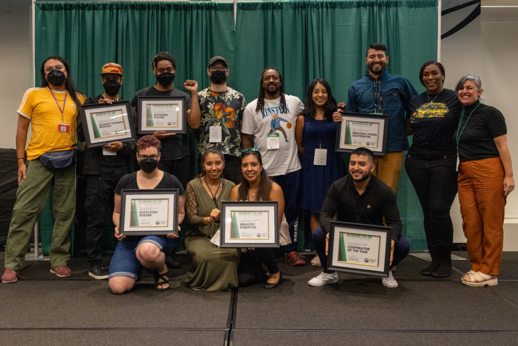 alrededor de doce personas se paran en dos filas en un escenario con certificados que dicen "worker co-ops awards" y sonrien