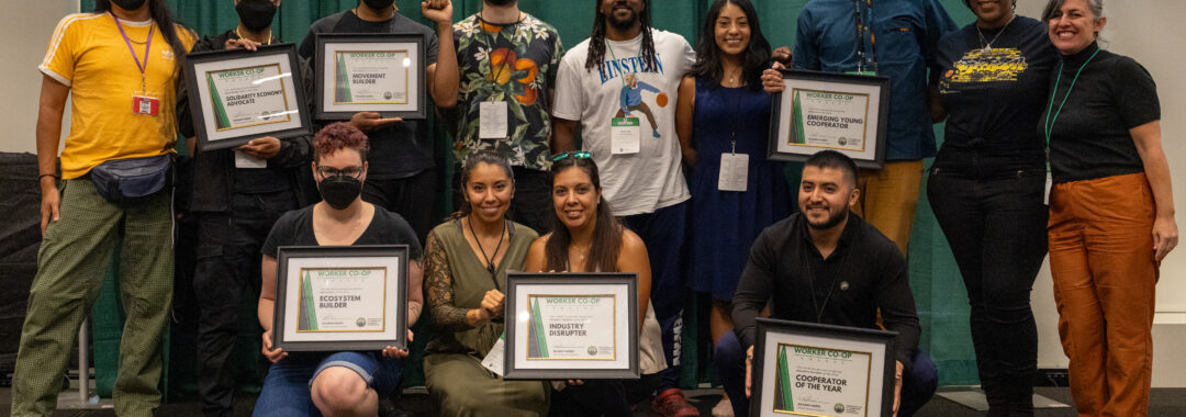 about twelve people stand in two rows on a stage holding certificates that say "worker co-op awards" and smile together