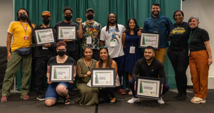 about twelve people stand in two rows on a stage holding certificates that say "worker co-op awards" and smile together