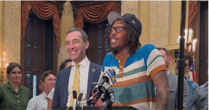 A photo of two people by a podium with a microphone smiling and looking off into a crowd outside the frame.