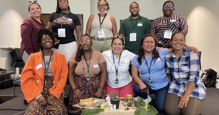 Around 10 people sit and stand together in a room with a small table in front of them with water, herbs and other offerings. The people wear lanyards and smile into the camera.
