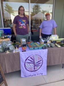 A photo of two people standing behind a table of produce with a sign that says yard farm co-op