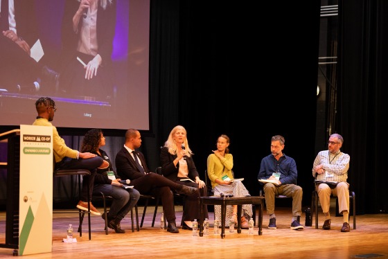 a group of six people sit in chairs on a stage and talk while a moderator sits in a stool and listens 